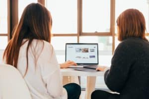 Two women from behind, side by side at a laptop in a bright window-lit office, screen showing a grid of images